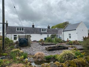A large white farm house sits in a garden of raised beds. A black oil tank is visible. The paths are gravelled and clean.