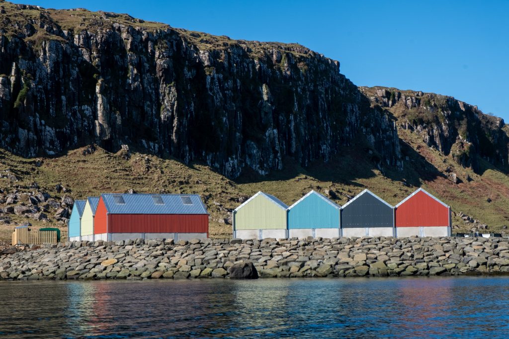 A group of brightly coloured industrial units on a harbour in front of dramatic cliffs.