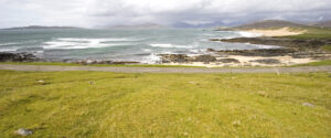 A coastline in West Harris - green grass is in the foreground with a road separating it from a beach with rocky outcrops. The sea is wavy and there is mountains in the background.