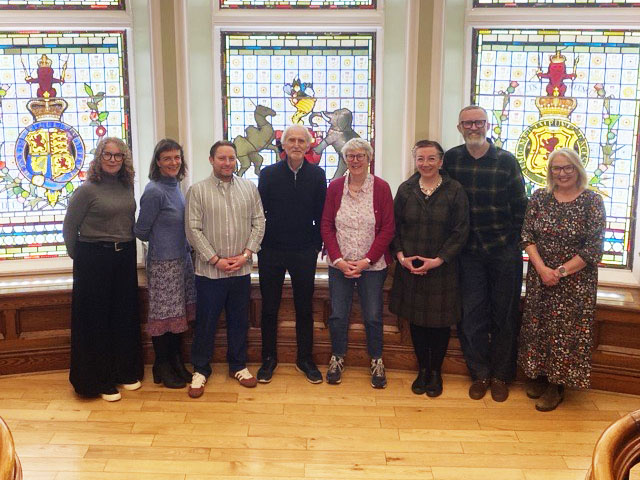 A group of eight people stand in front of three stained glass windows and smile for the camera