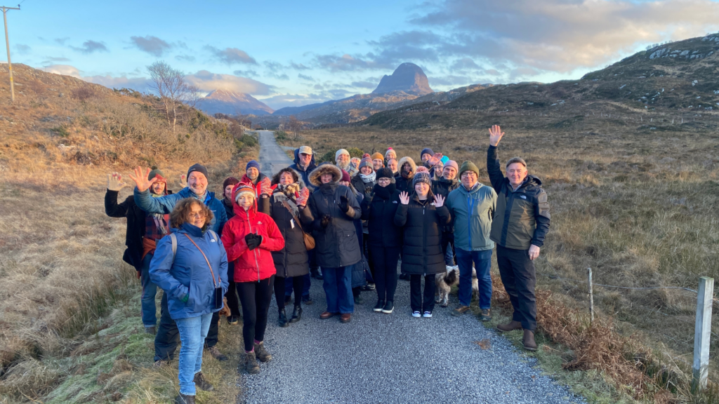 A group of people stand on a road in front of a mountain and wave to the camera