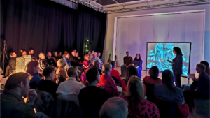 A group of people in a room listen to a person in front of a screen speak